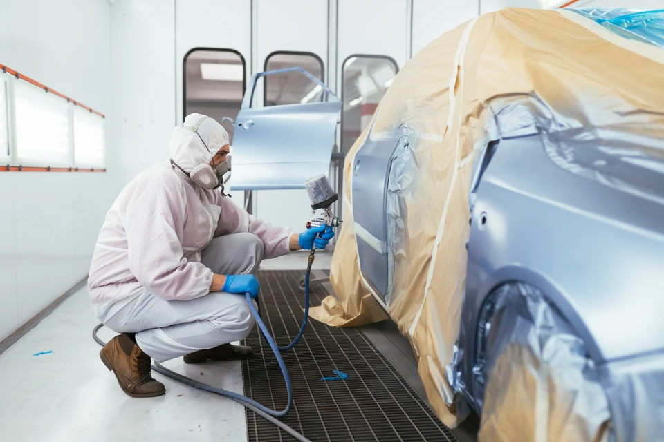 Atelier de carrosserie automobile avec technicien en combinaison de protection intégrale appliquant de la peinture au pistolet sur véhicule