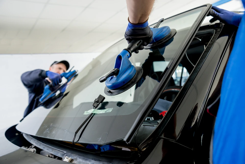 Scène d'atelier automobile en intérieur avec techniciens en combinaisons bleues effectuant le remplacement d'un pare-brise sur véhicule noir