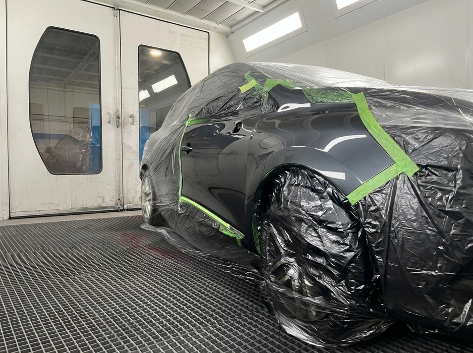 Carrosserie noire enveloppée de film de masquage plastique noir, maintenu par du ruban adhésif de masquage vert, dans une cabine de peinture professionnelle