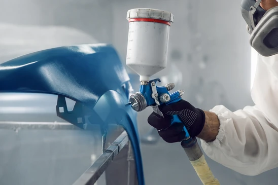 Atelier de carrosserie automobile avec technicien en combinaison de protection intégrale appliquant de la peinture au pistolet sur véhicule
